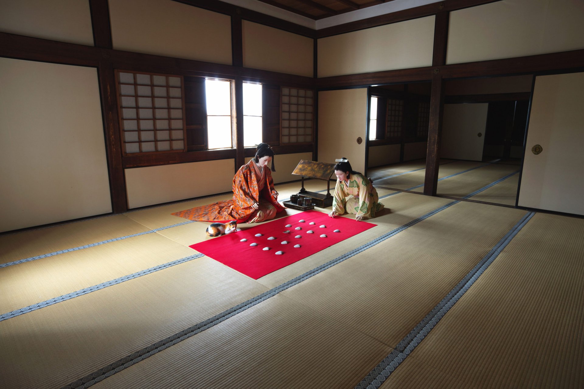 Two people in traditional kimonos sit on a red cloth playing a game inside a Japanese house, with a small cat nearby, captured in a serene HD photography scene.