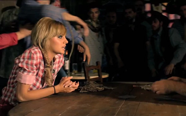 2K Quad HD PC desktop wallpaper: blonde pop singer in plaid shirt leaning on a wooden table, hands clasped, in a crowded dim music venue.