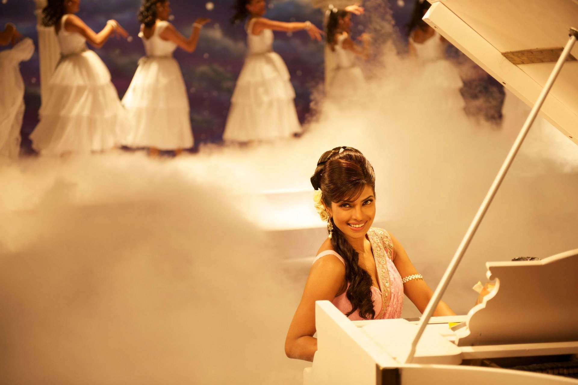 A vibrant HD desktop wallpaper featuring Priyanka Chopra sitting at a piano, surrounded by a misty atmosphere and dancers in white dresses, exuding elegance and charm.