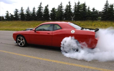 A vibrant red tuned vehicle performs a burnout, releasing thick smoke as it accelerates on an empty road, framed by lush green trees in the background. Perfect HD desktop wallpaper.