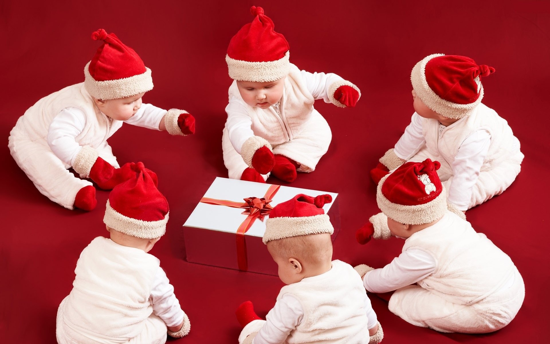 A group of cute babies in red Santa hats sit on a festive red background, joyfully gathered around a beautifully wrapped Christmas gift, celebrating the holiday spirit.