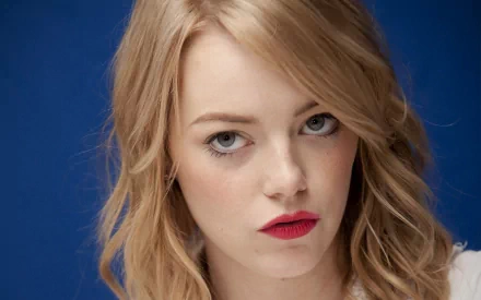 HD desktop wallpaper featuring a close-up portrait of celebrity Emma Stone with natural makeup and red lipstick against a solid blue background.