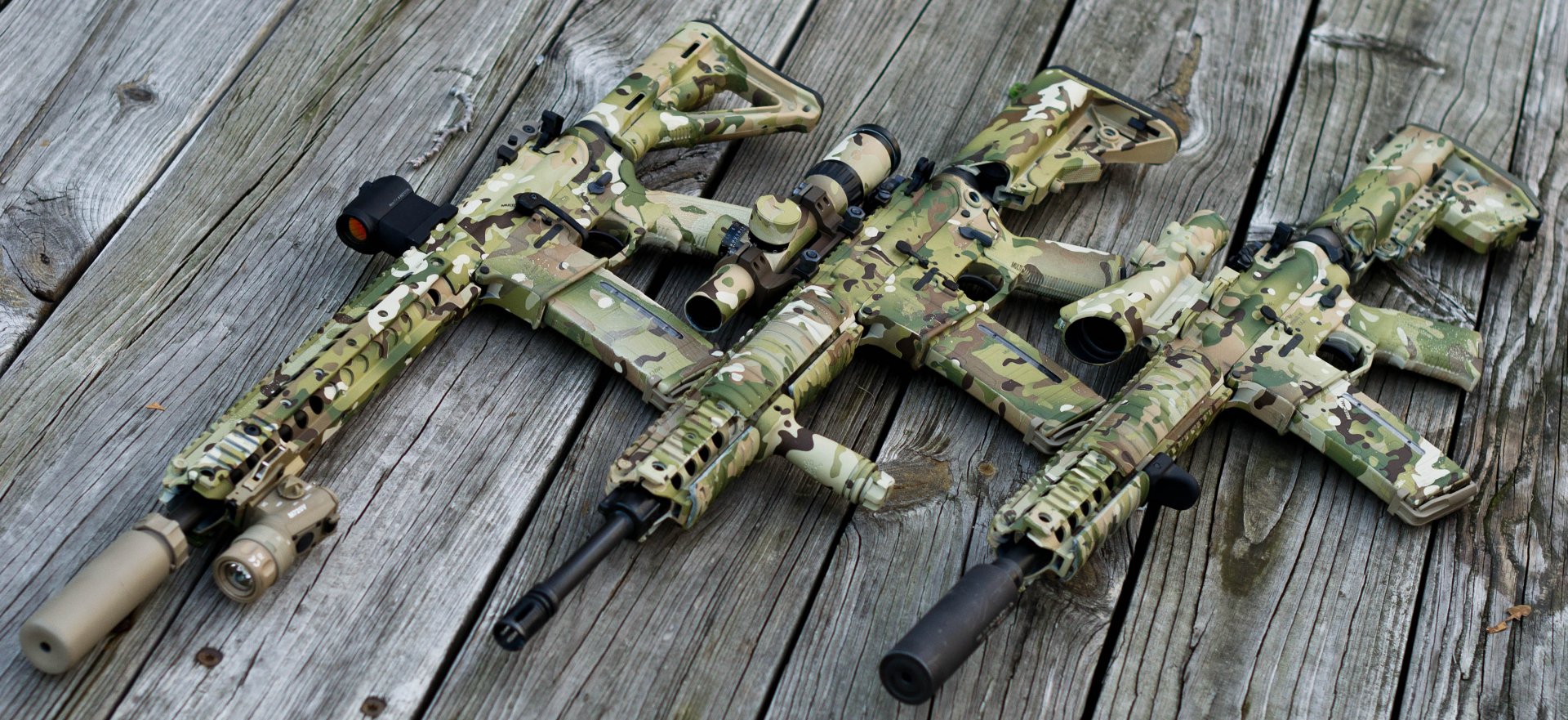 Man-made camouflaged assault rifles laid on weathered wooden planks — 2K Quad HD PC desktop wallpaper/background.
