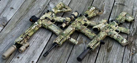 Man-made camouflaged assault rifles laid on weathered wooden planks — 2K Quad HD PC desktop wallpaper/background.