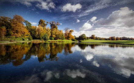 HD desktop wallpaper featuring a beautiful fall landscape with vibrant trees reflected in the calm lake water under a partly cloudy sky.