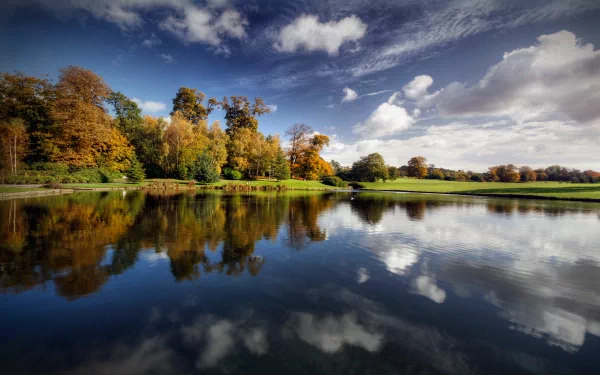HD desktop wallpaper featuring a beautiful fall landscape with vibrant trees reflected in the calm lake water under a partly cloudy sky.