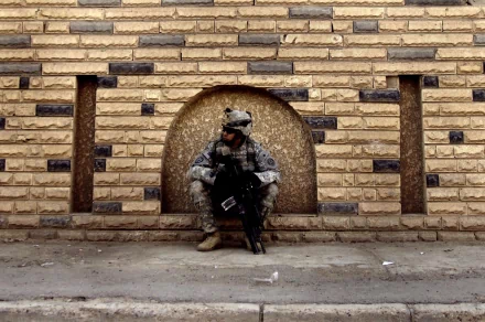 HD wallpaper showing a soldier in military gear, sitting against a brick wall, holding a rifle. Background features tan-colored bricks with an arched inset, providing a rugged, urban setting.