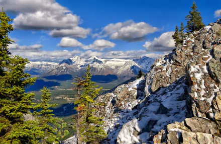 A breathtaking HD desktop wallpaper showcasing a mountain landscape, featuring rocky terrain, snowy peaks, and lush green trees under a vibrant blue sky with fluffy clouds.