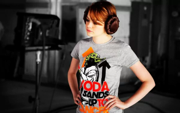 Emma Stone poses in a grey T-shirt featuring Yoda, wearing headphones, in a creative setting. This high-definition image serves as a vibrant desktop wallpaper.
