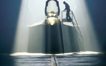 A silhouette of a technician working on a Lockheed Martin F-22 Raptor, illuminated by shafts of light in a hangar, showcasing the aircraft's sleek design.