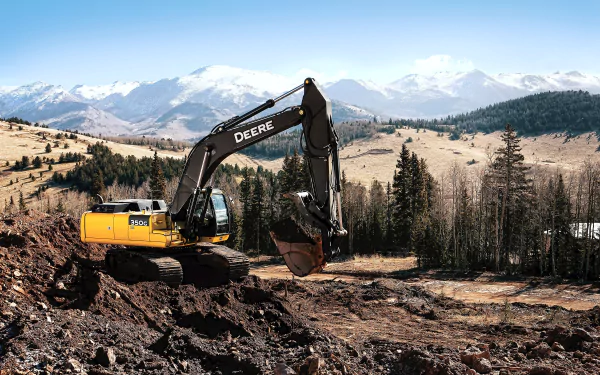 2K Quad HD PC desktop wallpaper: excavator vehicle digging on a mountain slope with pine forest and snow-capped peaks beneath a clear blue sky.