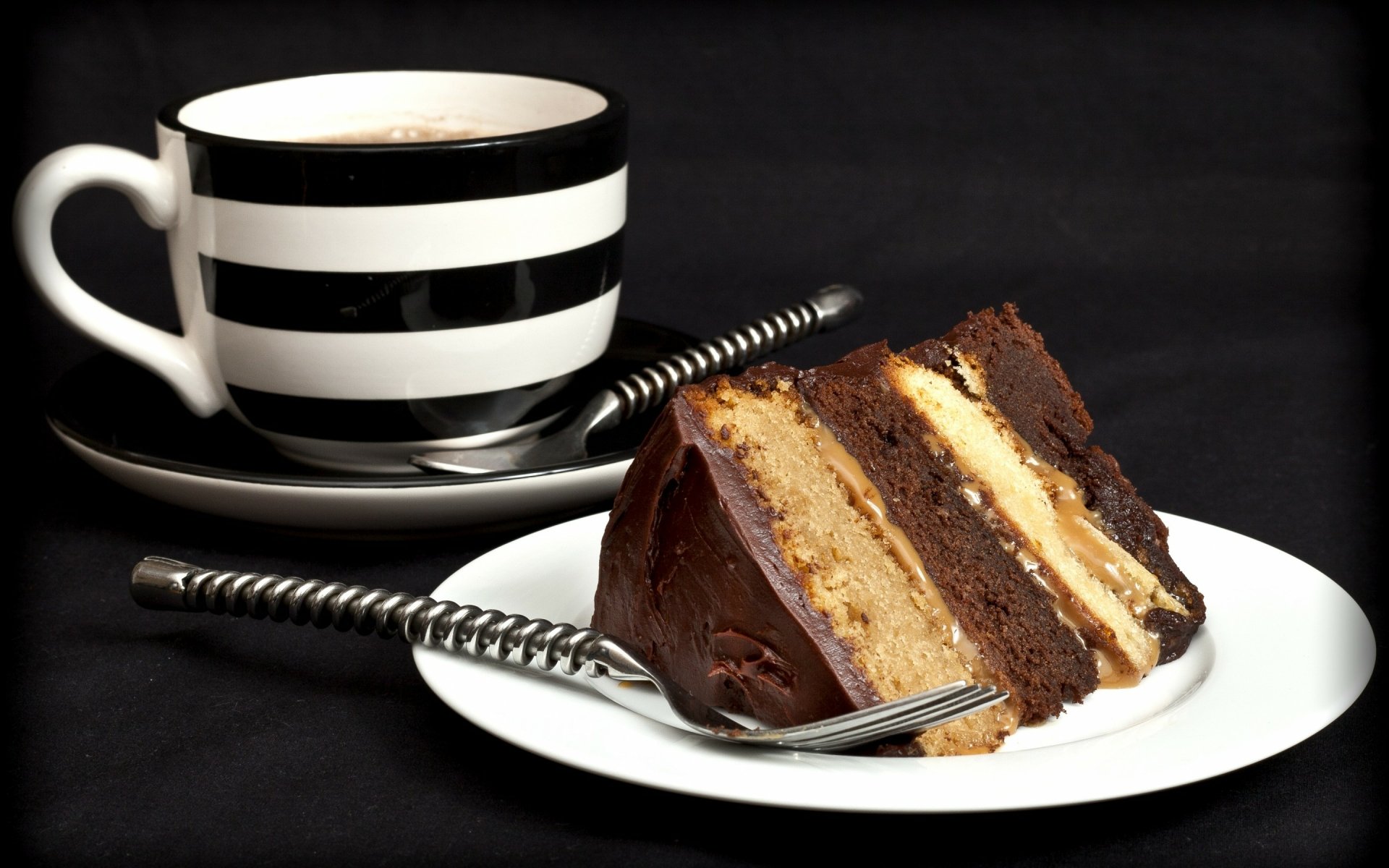 A decadent slice of chocolate cake layered with cream sits on a plate, accompanied by a black and white striped coffee cup, creating an appetizing food-themed HD desktop wallpaper.