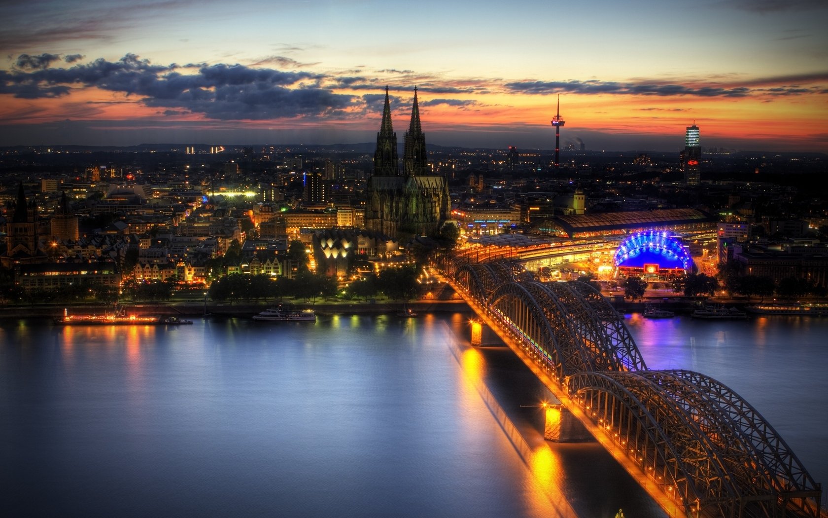 HD desktop wallpaper showcasing a vibrant nighttime view of Cologne with illuminated cityscape, the cathedral, and the glowing Hohenzollern Bridge over the river.