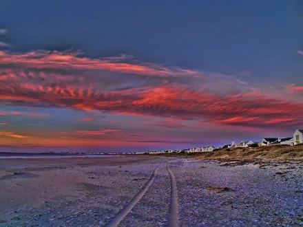 HD desktop wallpaper: Cape York coastline at sunset — vibrant pink-orange clouds over a sandy beach with tire tracks and low coastal cottages.