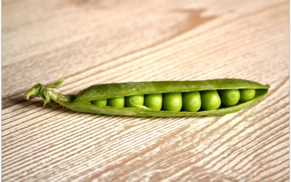 HD desktop wallpaper featuring a fresh green pea pod opened to reveal a row of vibrant peas on a light wooden surface.