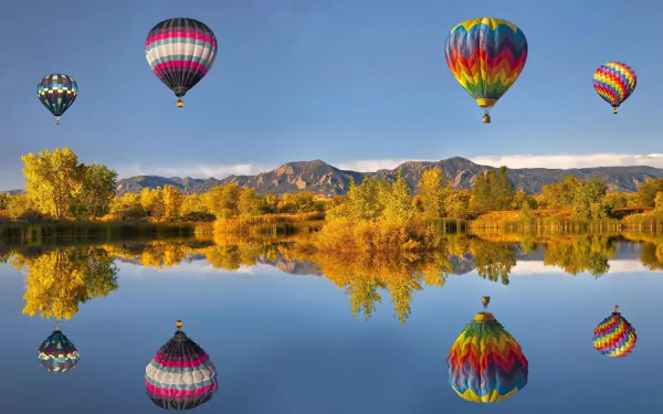 HD wallpaper of colorful hot air balloons floating above a serene lake with mirrored reflections, set against a backdrop of mountains and autumn trees.