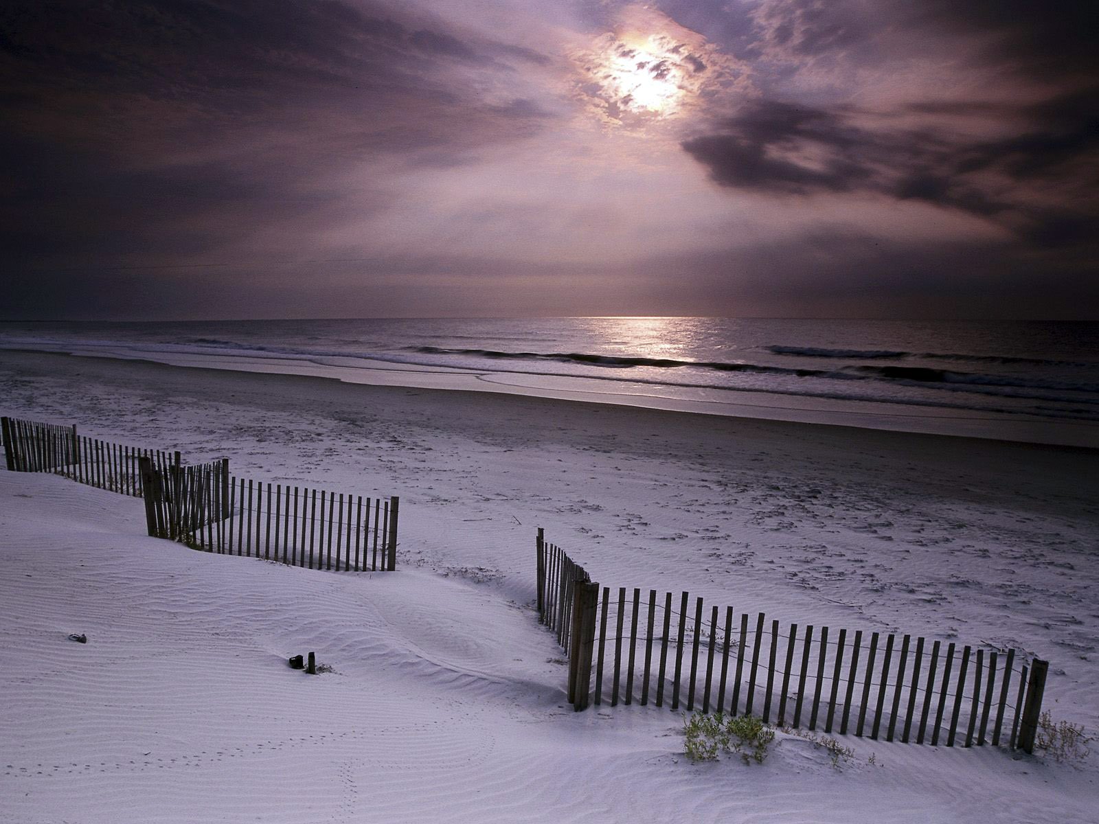 HD desktop wallpaper of a serene beach at dusk, featuring calm waves, soft sand, and wooden fences under a cloudy sky with the moonlight reflecting on the water.