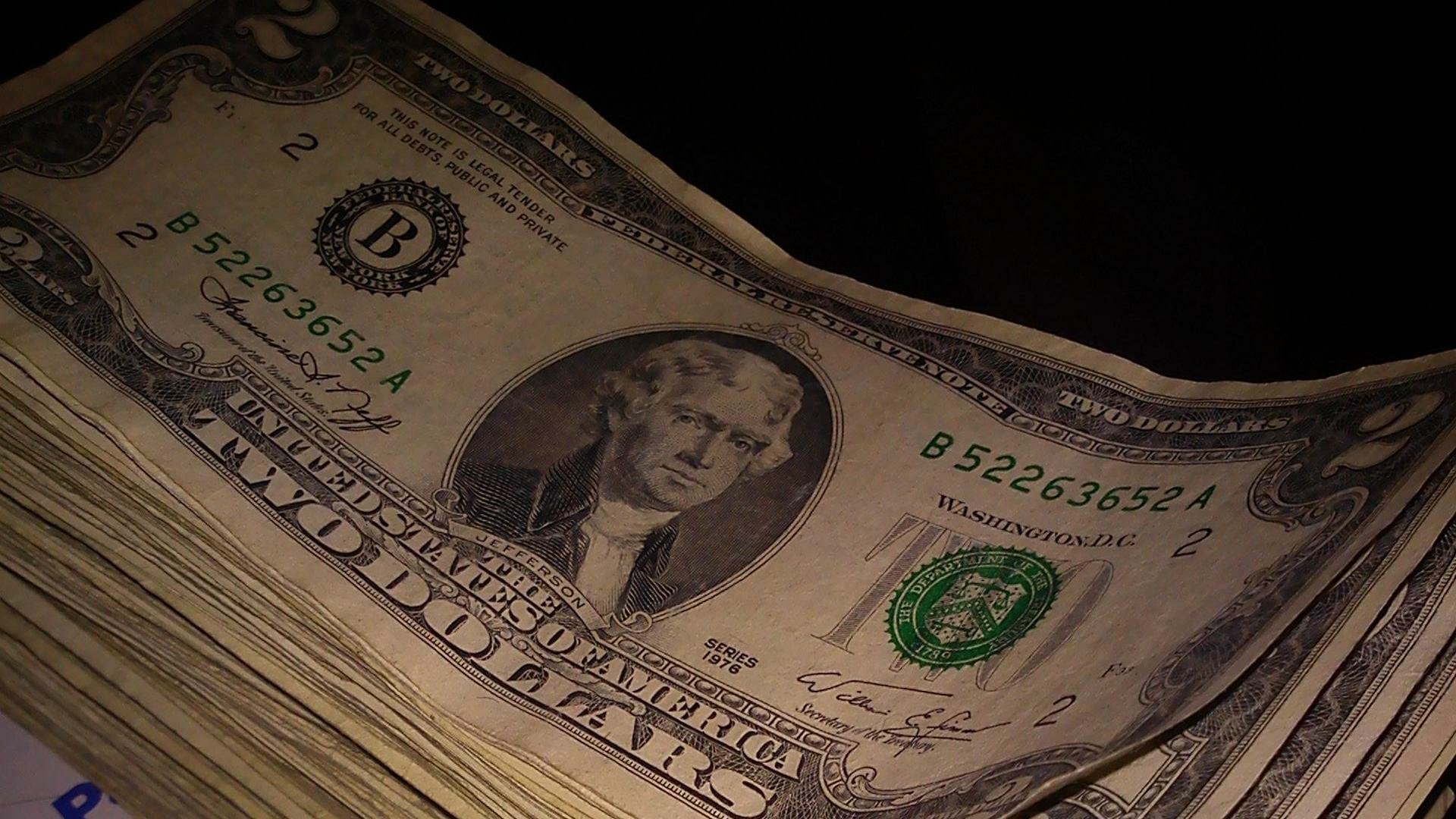 HD PC desktop wallpaper showing a man-made stack of US dollar bills, close-up on a worn portrait, green seals and serial numbers visible against a dark background.