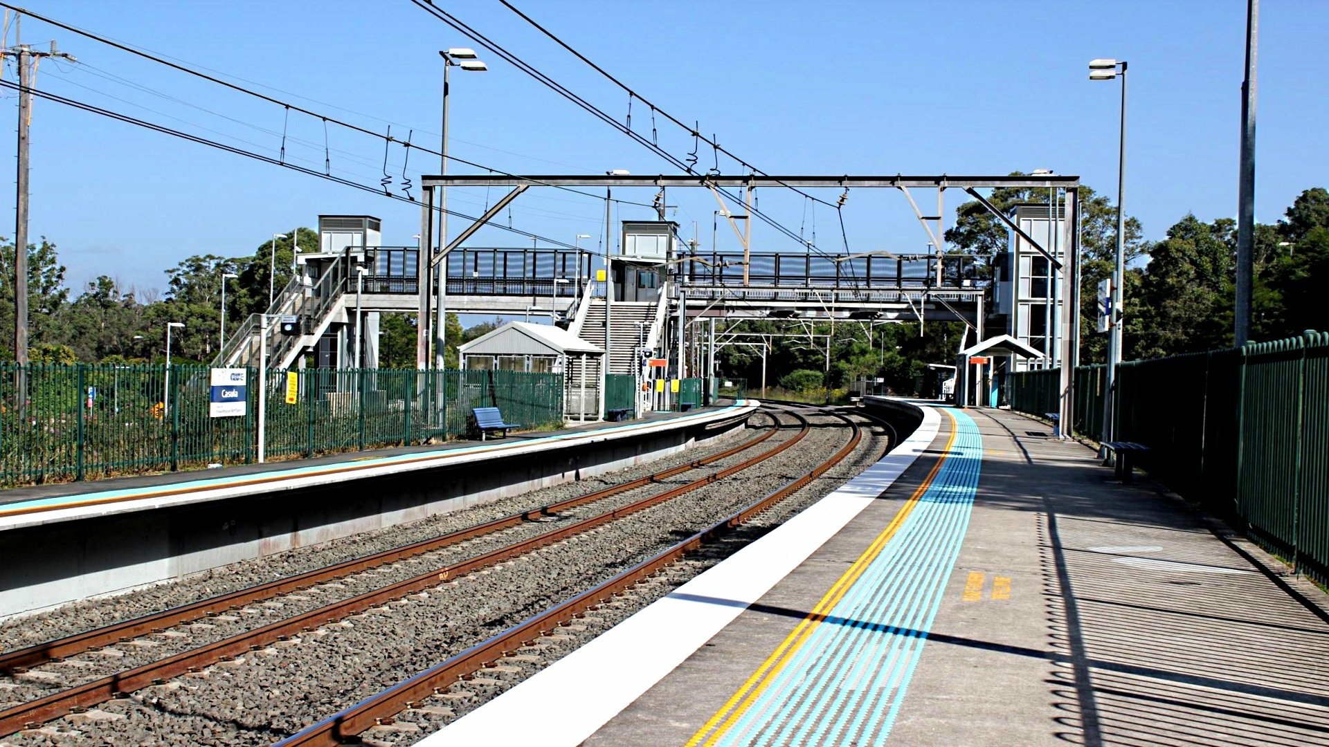 An HD desktop wallpaper and background of a man-made train station featuring empty platforms, railway tracks, overhead wires, and a pedestrian bridge under clear blue skies.