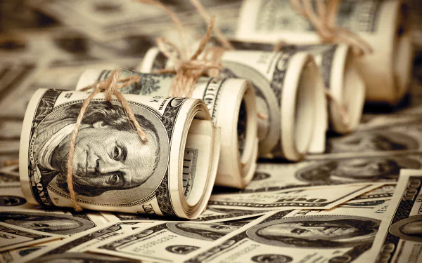 A close-up of rolled bundles of dollar bills, neatly tied with twine, set against a background of scattered dollar notes, creating an engaging man-made wallpaper design.