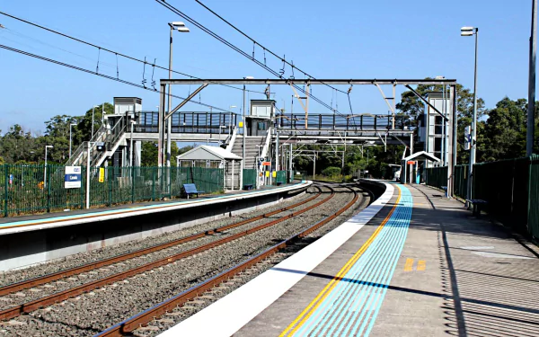 An HD desktop wallpaper and background of a man-made train station featuring empty platforms, railway tracks, overhead wires, and a pedestrian bridge under clear blue skies.
