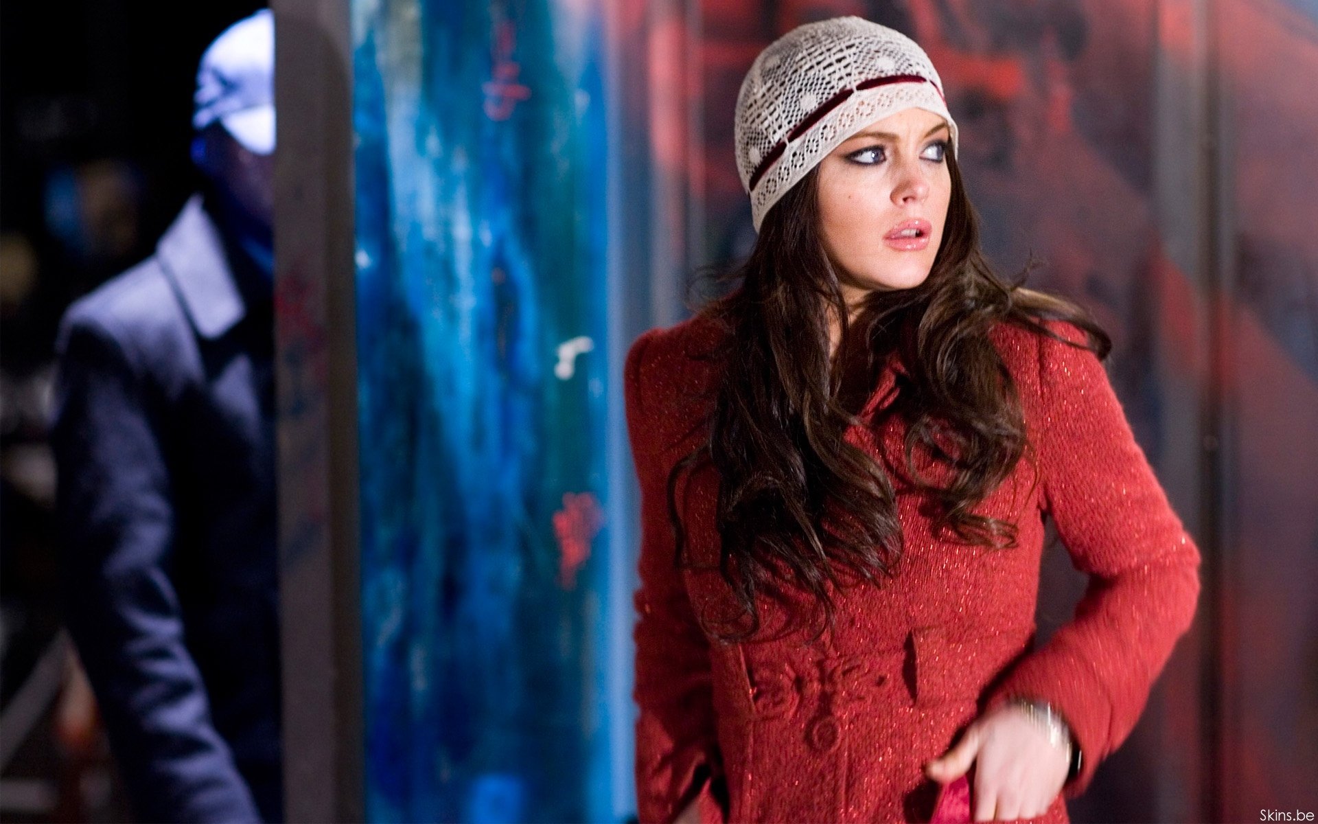 HD PC desktop wallpaper of a celebrity woman in a red coat and knit beanie, gazing right against a blurred, colorful urban backdrop.