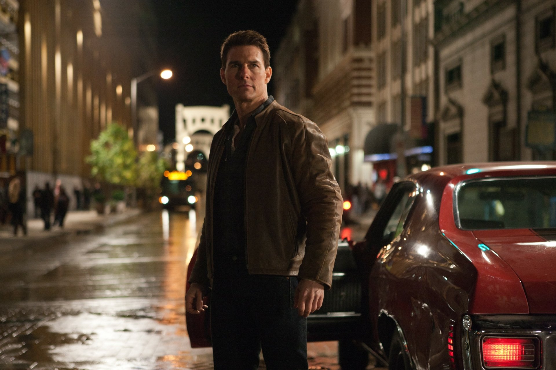 2K Quad HD PC desktop wallpaper: movie-style image of a man in a brown jacket standing by a red car on a wet city street at night, tense thriller atmosphere.