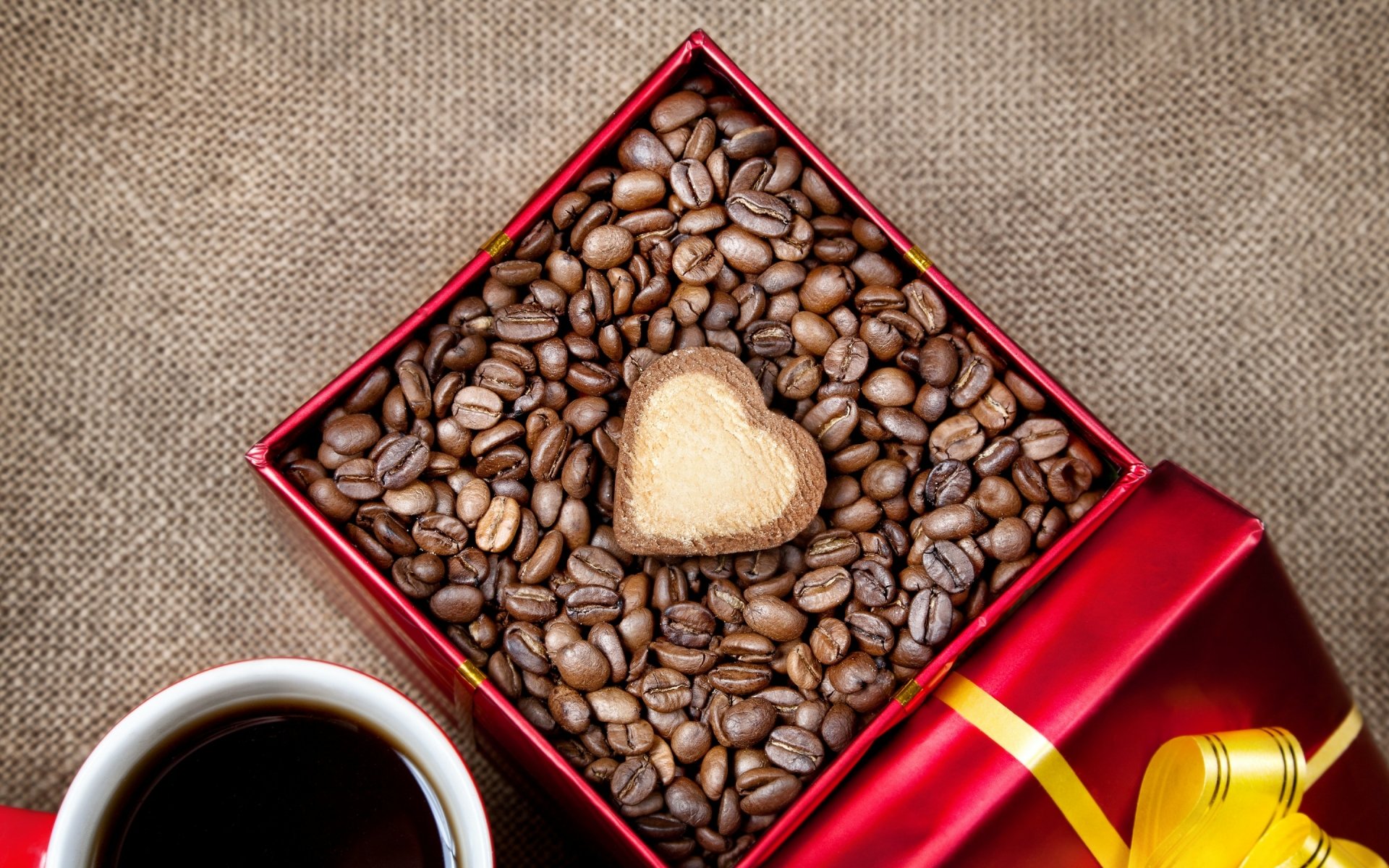 A rich arrangement of coffee beans in a red gift box, featuring a heart-shaped chocolate on top, accompanied by a steaming cup of coffee, creating a warm, inviting scene.
