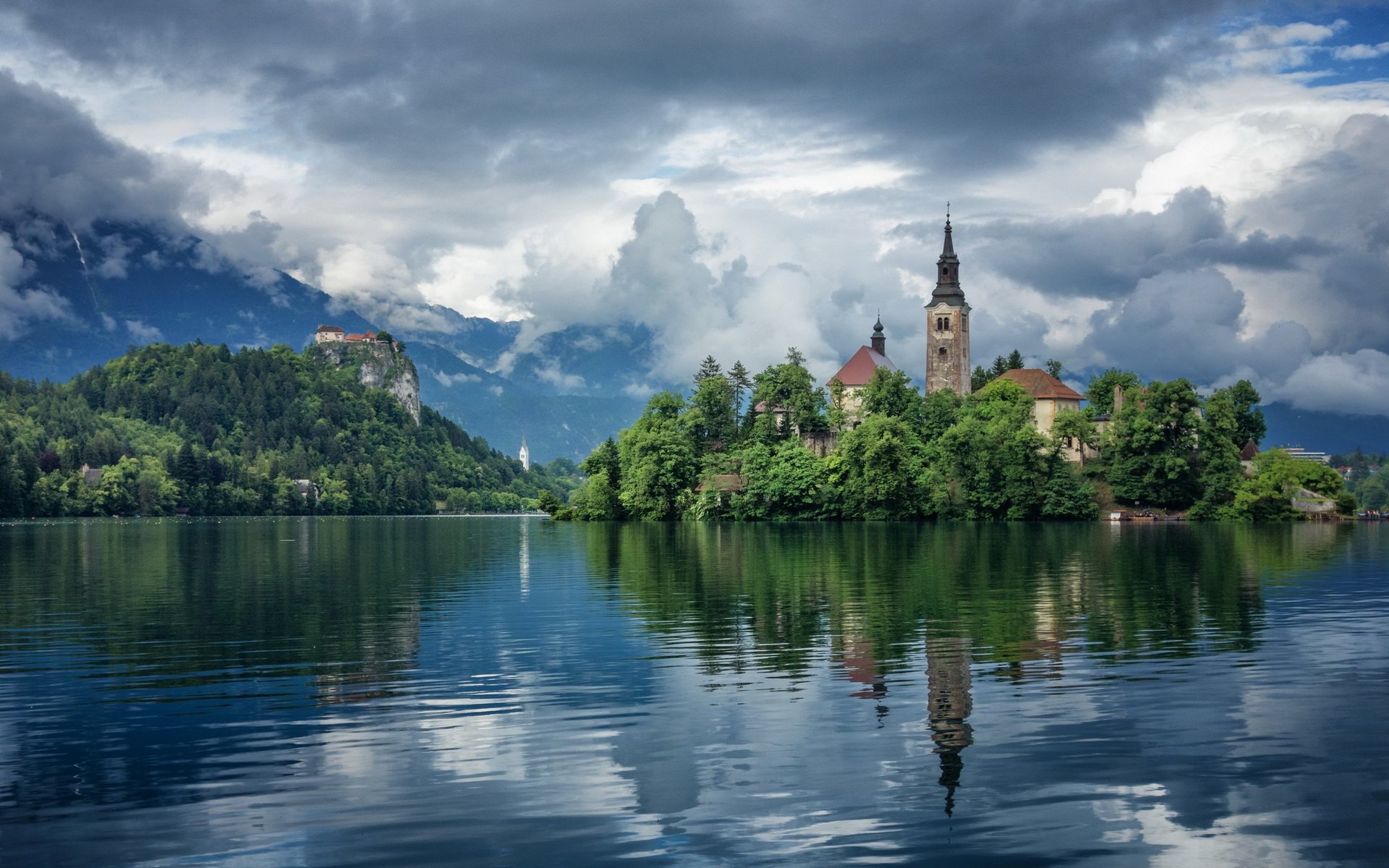A tranquil scene of the Pilgrimage Church of the Assumption of Maria in Slovenia, surrounded by lush greenery and reflected in the serene waters of Lake Bled under a dramatic sky.