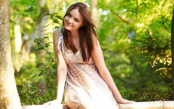 A woman in a light dress smiles while sitting on a wooden fence, surrounded by lush green trees in a serene outdoor setting. HD desktop wallpaper and background.