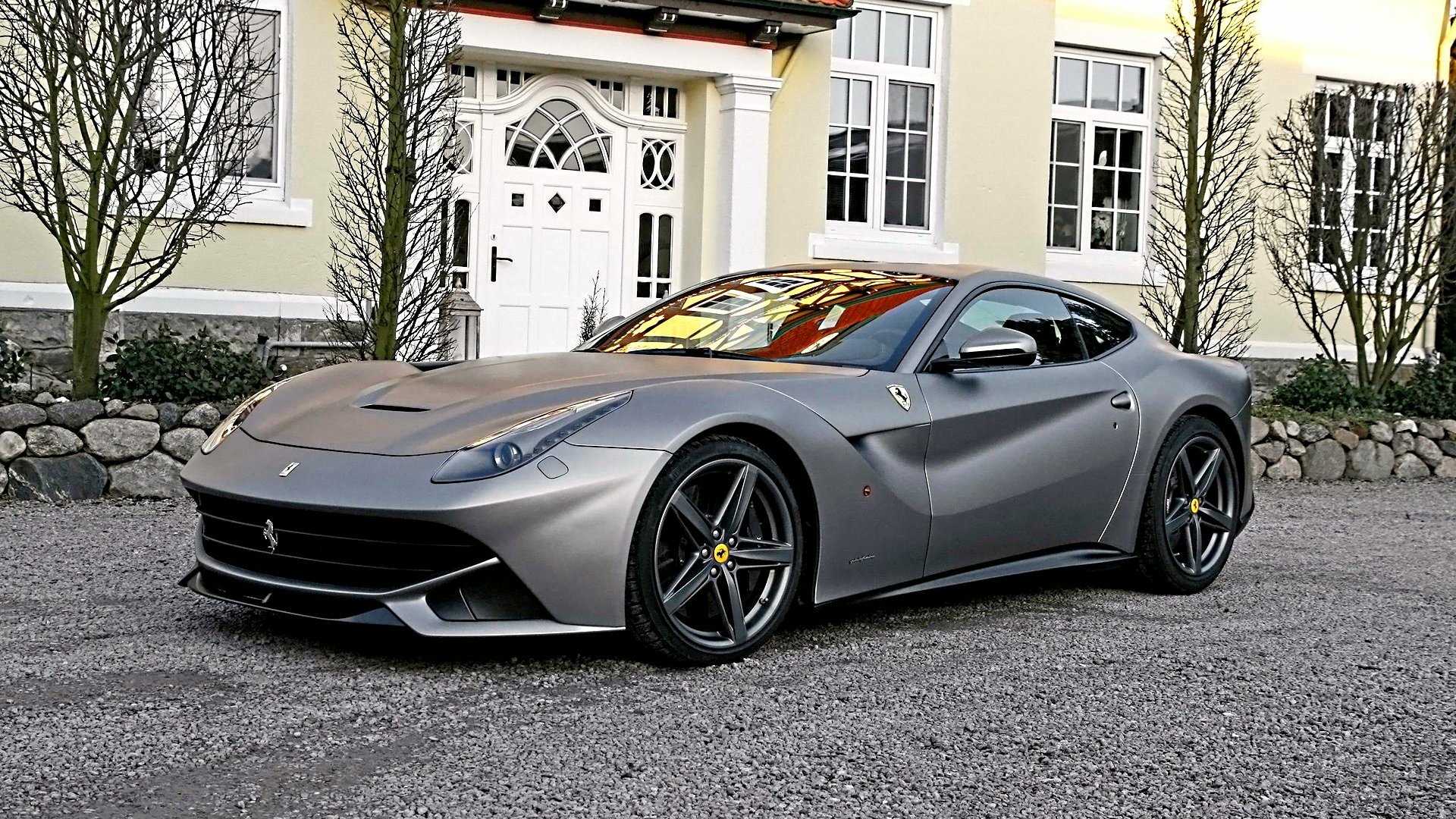 HD PC desktop wallpaper showing a silver Ferrari F12berlinetta vehicle parked in front of a house, three-quarter front view emphasizing sleek curves and alloy wheels.