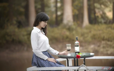 HD desktop wallpaper of an Asian woman sitting at an outdoor table near a pond, with a bottle and glass of wine, a red flower, and a blurred forest background.