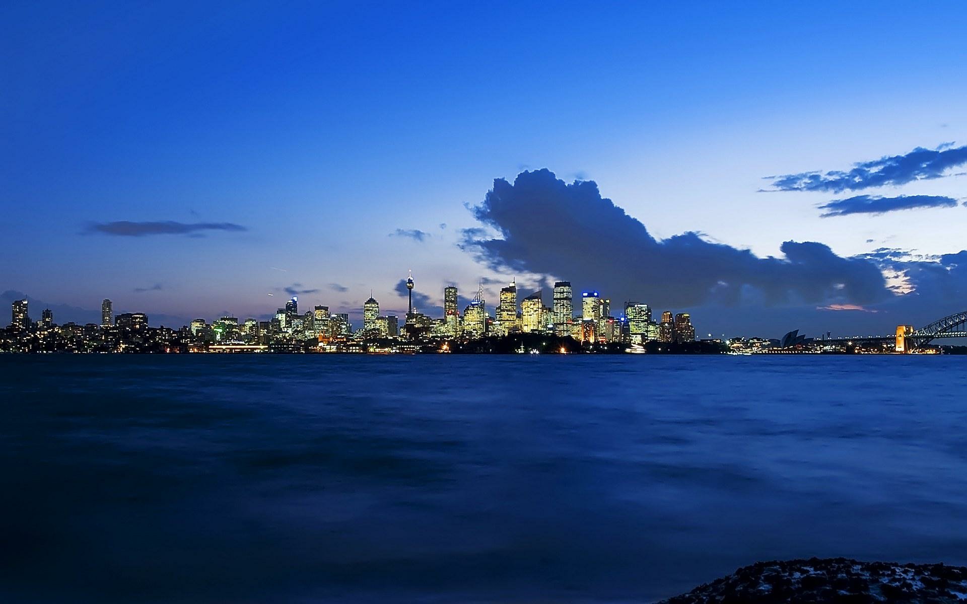 HD PC desktop wallpaper: man-made Sydney skyline at dusk across the harbor, illuminated towers and waterfront lights reflecting on calm water.
