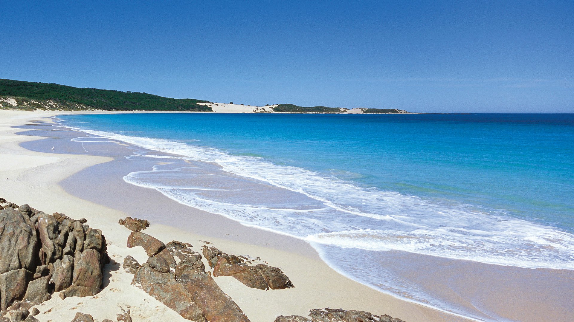 HD PC desktop wallpaper showcasing a serene beach with clear blue water, gentle waves, white sand, and a natural coastline under a bright blue sky.