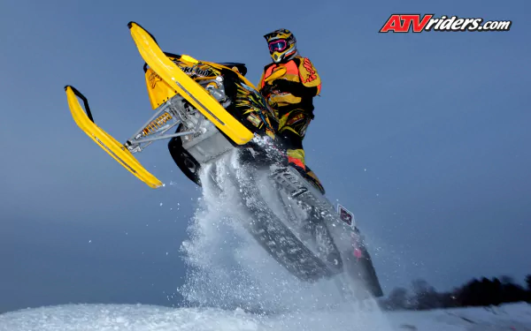 HD PC desktop wallpaper: yellow snowmobile and rider launching off a snowy jump against a clear blue sky.