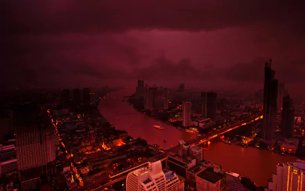 A dramatic view of Bangkok at night, showcasing the illuminated skyline and river under a moody, dark sky. This HD desktop wallpaper captures the city's vibrant urban landscape.