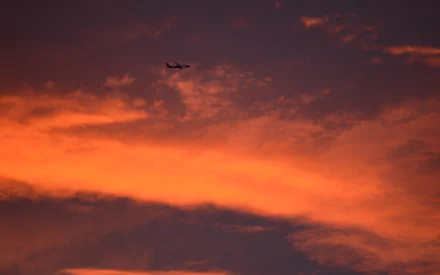 An airplane glides through a vibrant sunset sky filled with dramatic clouds, capturing the beauty of nature alongside the contrast of a distant bushfire.