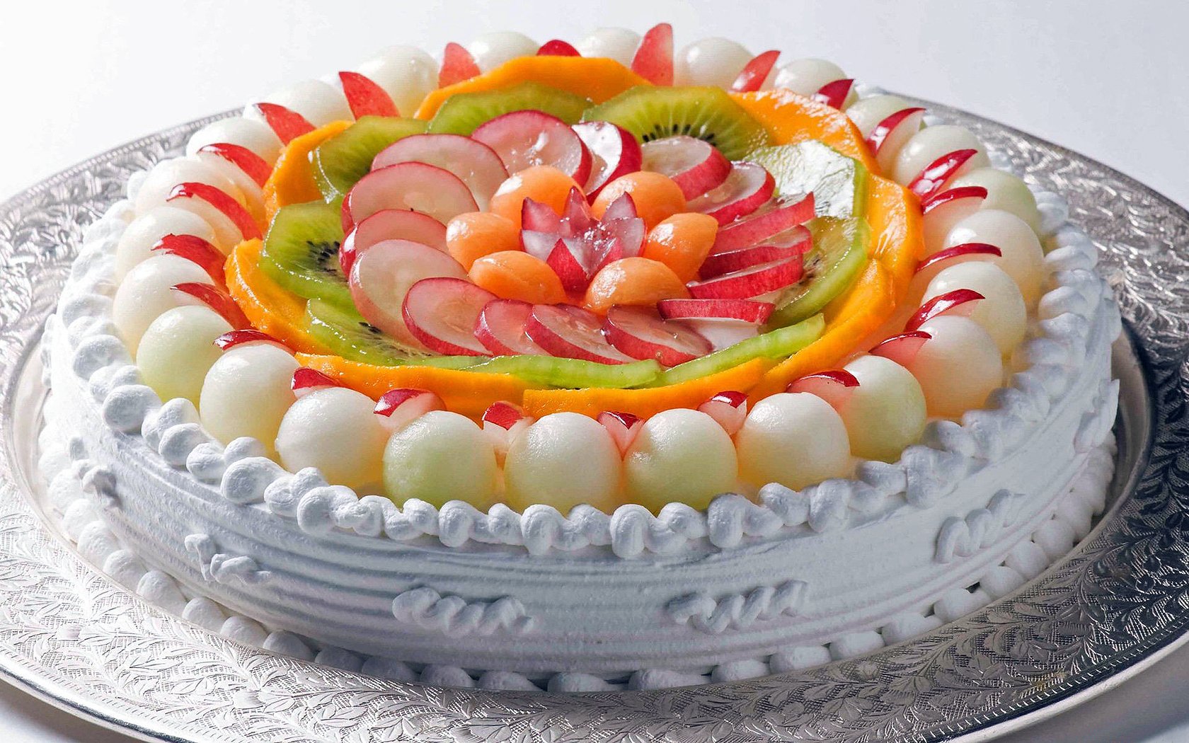 A beautifully decorative fruit cake, topped with a variety of colorful fruits arranged in a stunning floral design, set against a silver plate background. A vibrant HD desktop wallpaper.