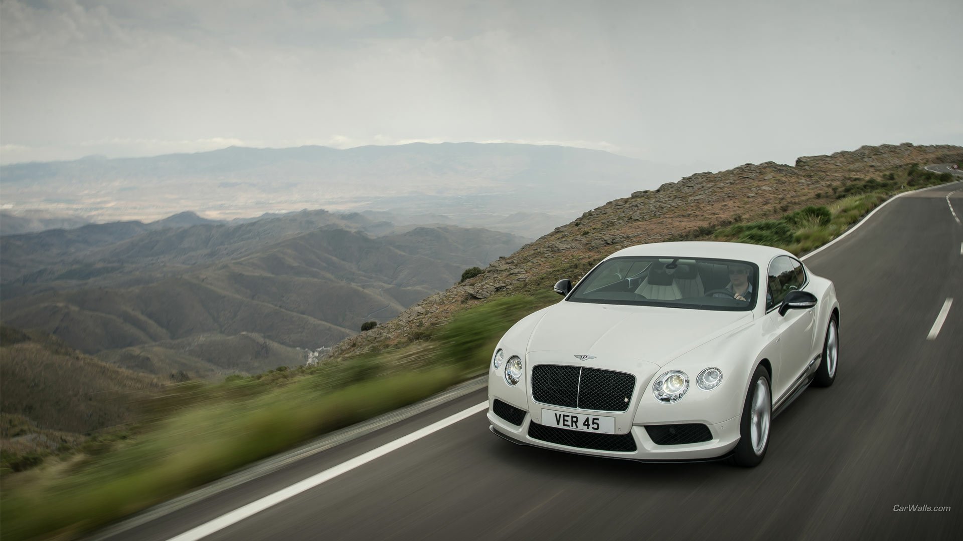 White Bentley Continental GT V8 cruising a winding mountain road with sweeping valley vistas — HD PC desktop wallpaper background.