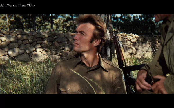 HD PC desktop wallpaper/background from the movie Kelly's Heroes: a World War II soldier in olive uniform sits by a stone wall with a rifle, gazing upward in sunlit grass.
