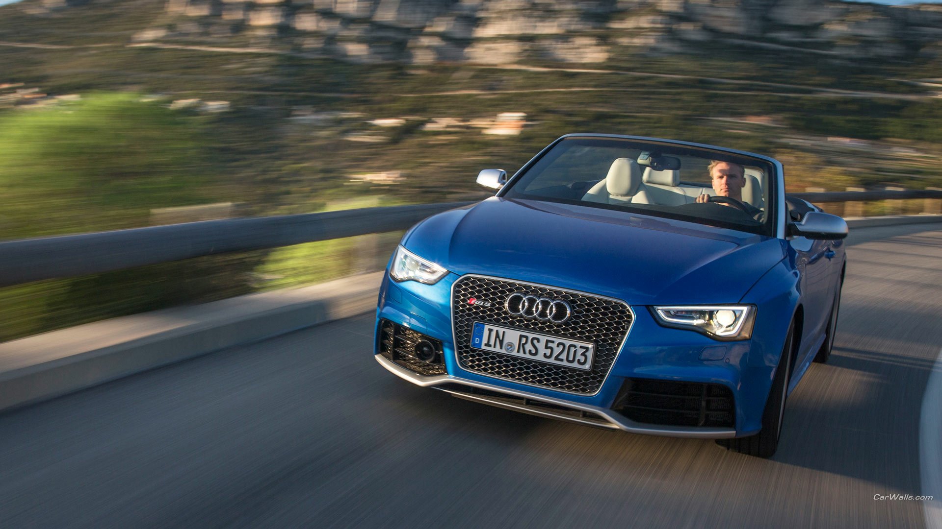 HD PC desktop wallpaper: blue Audi RS5 convertible speeding along a coastal road, driver visible, motion blur conveys dynamic performance.