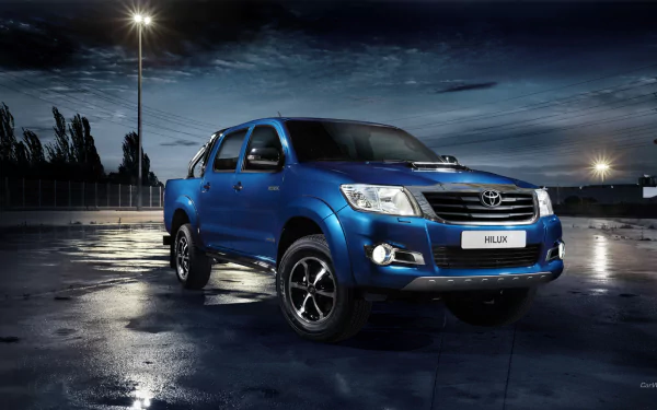 HD desktop wallpaper featuring a blue Toyota Hilux pickup truck parked on a wet surface under streetlights at dusk.