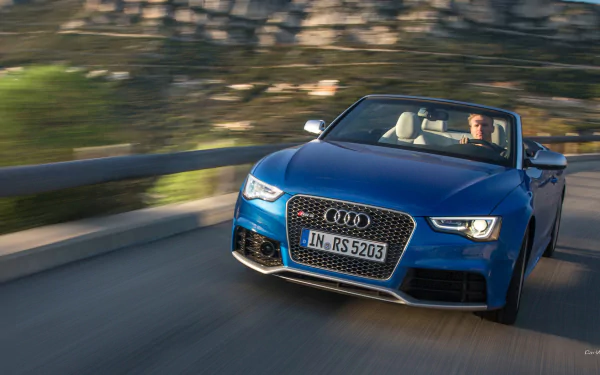 HD PC desktop wallpaper: blue Audi RS5 convertible speeding along a coastal road, driver visible, motion blur conveys dynamic performance.
