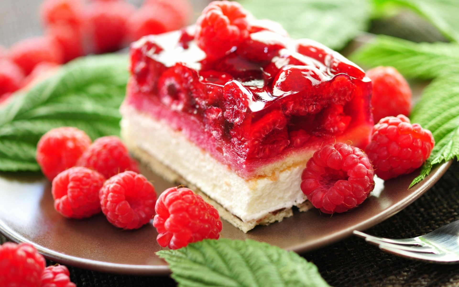 HD desktop wallpaper featuring a close-up of a vibrant raspberry-topped layered cake garnished with fresh raspberries and green leaves on a dark plate.