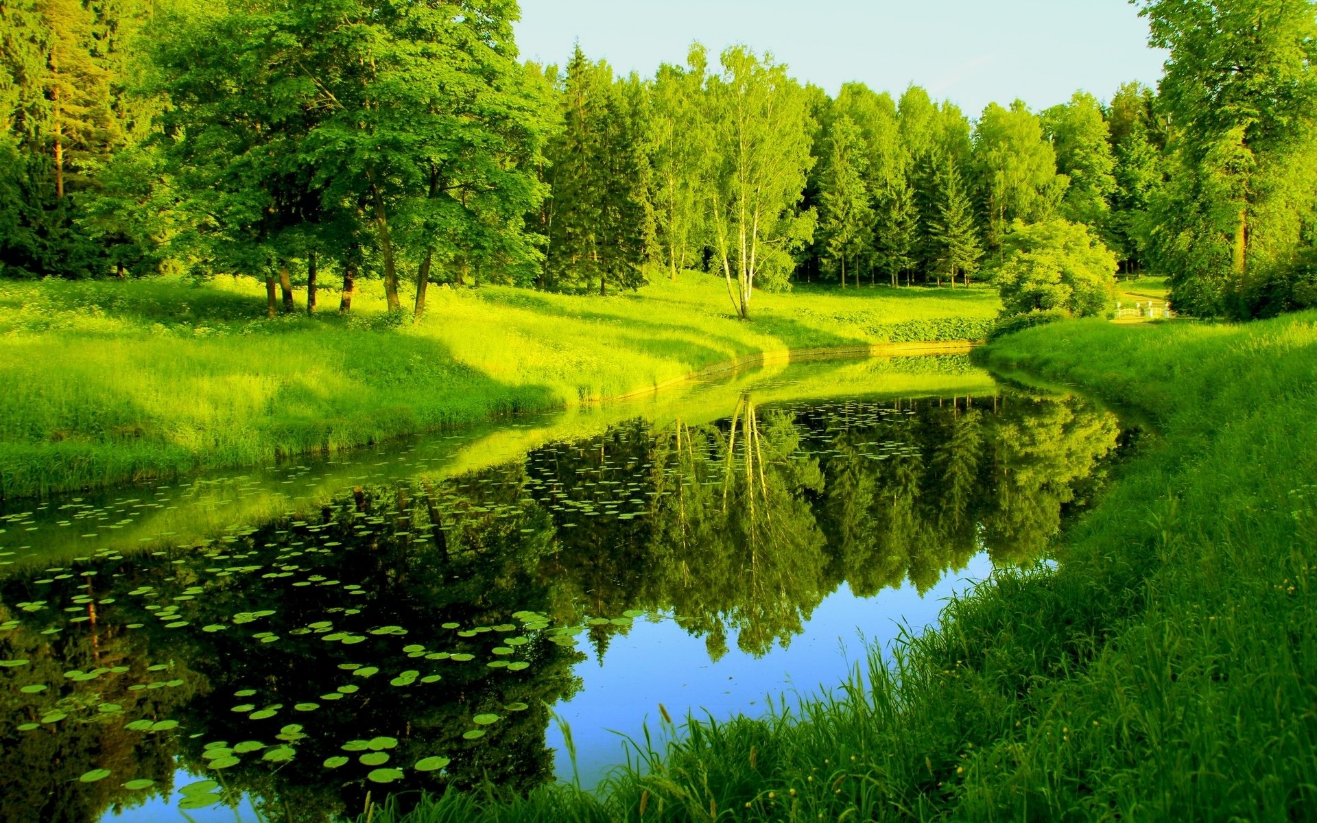 HD PC desktop wallpaper of a serene nature scene featuring a calm river reflecting lush green trees and grass under a clear sky.