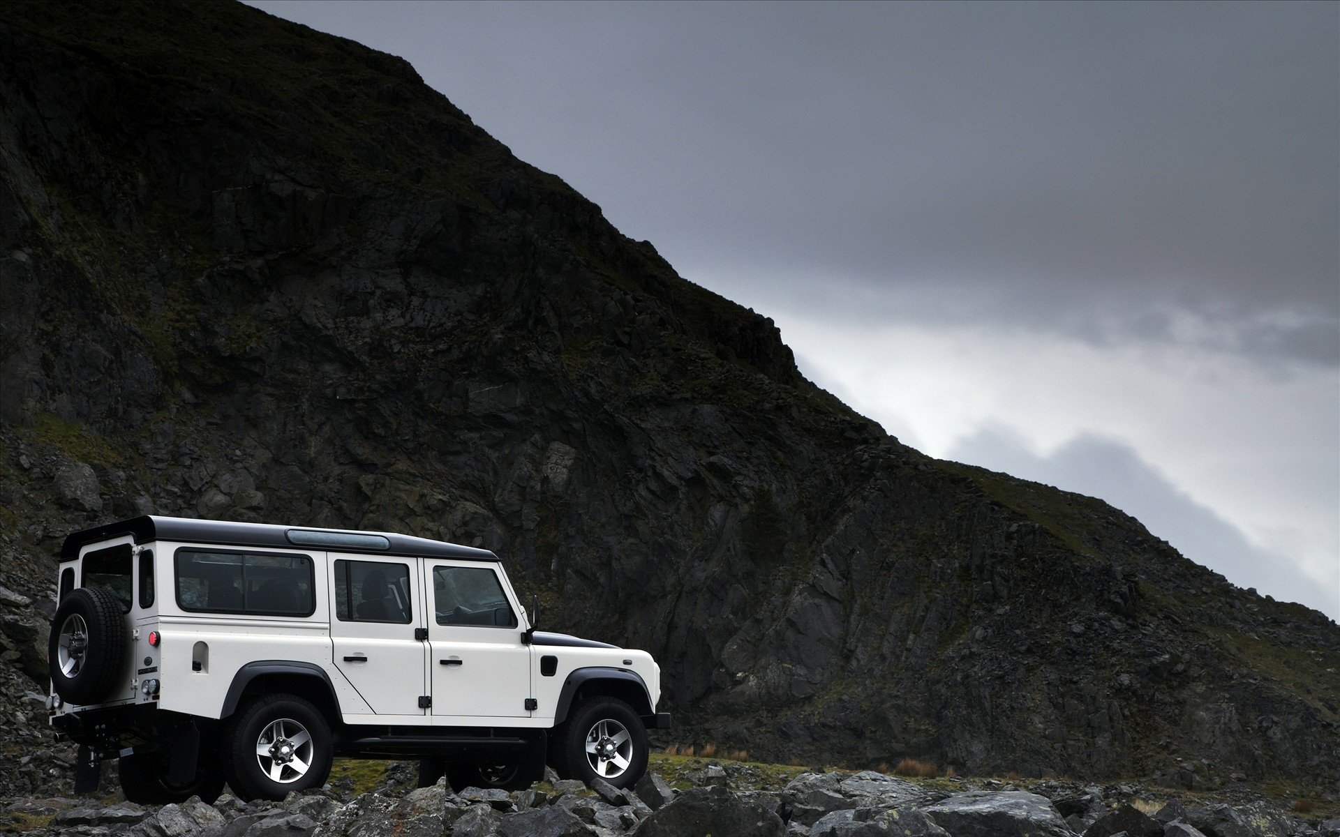 White Land Rover on a rocky slope beneath a stormy sky, dramatic mountain backdrop — HD PC desktop wallpaper/background.