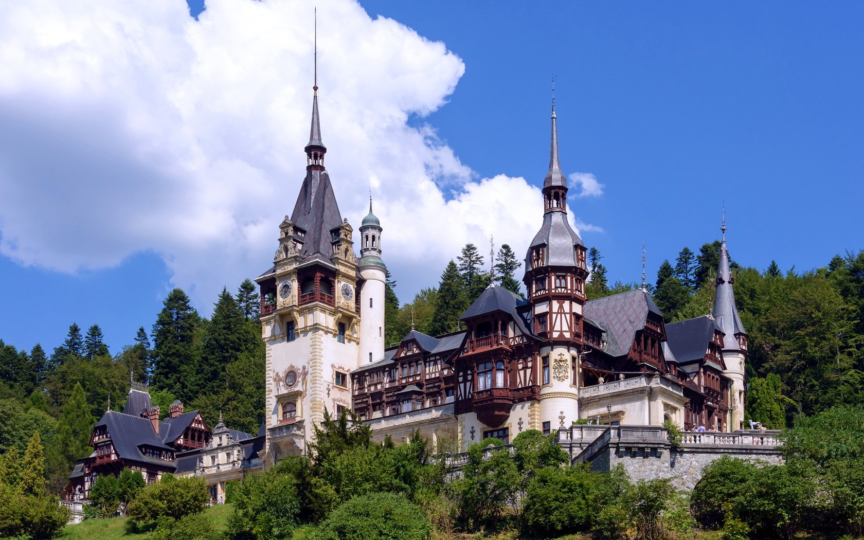 HD desktop wallpaper of Peles Castle, a beautifully detailed man-made structure set against a bright blue sky with lush greenery surrounding it.