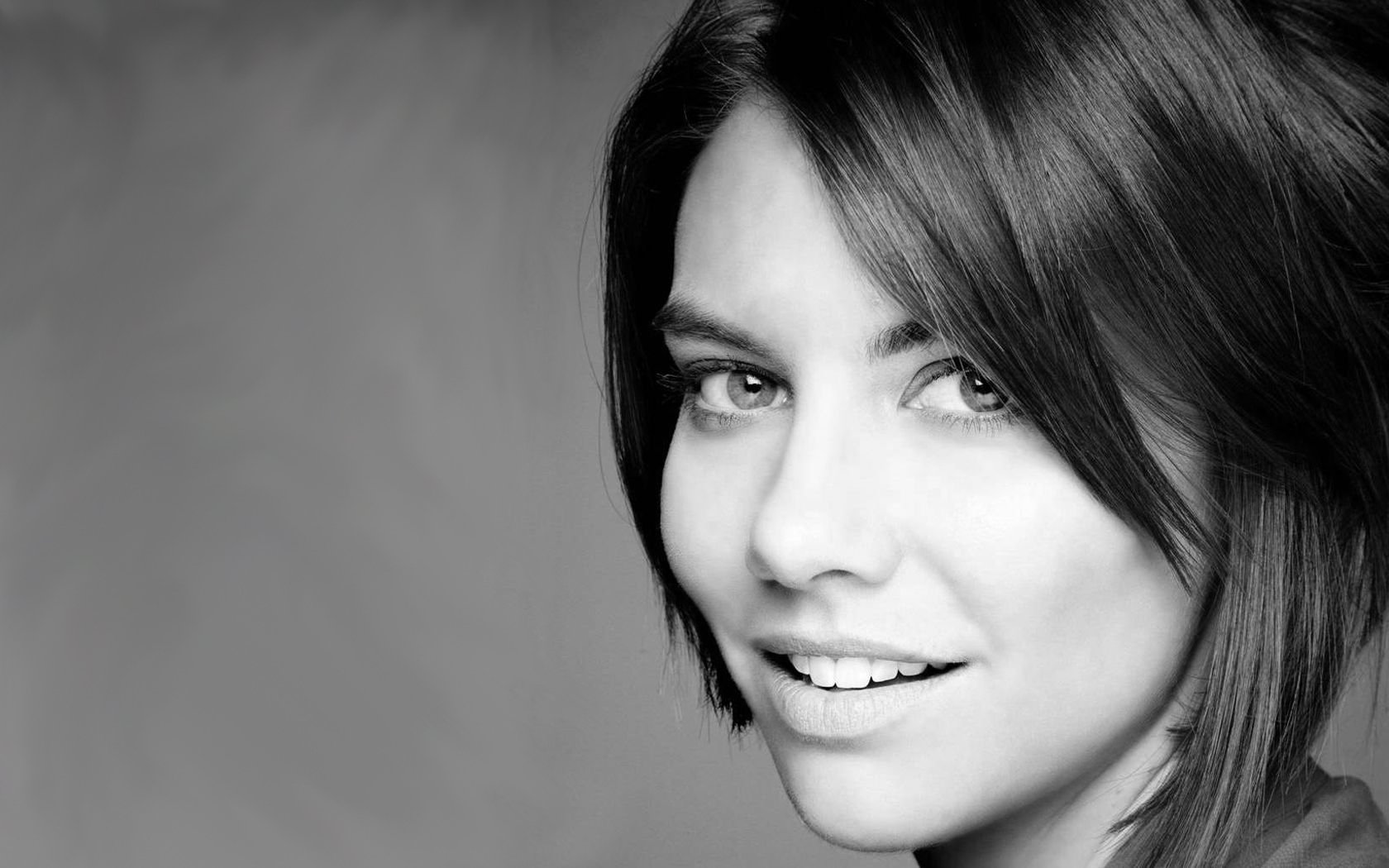 Black and white HD desktop wallpaper featuring a portrait of celebrity Lauren Cohan, showcasing her captivating smile and expressive eyes against a smooth background.