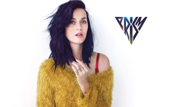 A high-definition desktop wallpaper featuring Katy Perry with dark hair, wearing a fuzzy yellow sweater, alongside the PRISM logo.