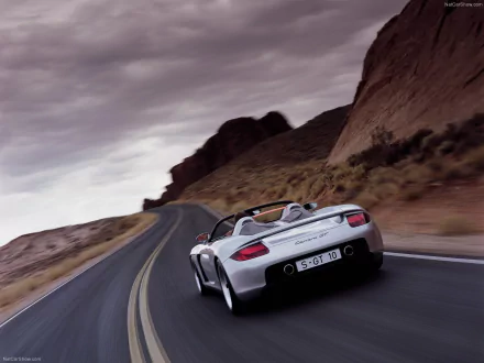 A sleek Porsche Carrera GT speeds along a winding mountain road, showcasing its dynamic design against a dramatic backdrop of rocky terrain and moody skies.